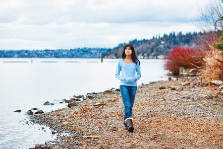 Young biracial teen girl in blue shirt and jeans walking along rocky shoreline of lake in early spring or fallの写真素材