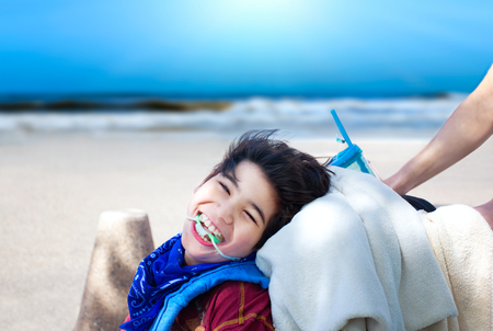 Happy disabled boy being pushed in wheelchair, smiling,  with ocean beach in backgroundの写真素材