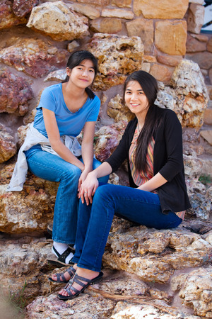 Two biracial  young teen girls, sisters or friends,  sitting together on rocky stone seats outdoorsの写真素材