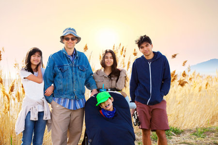 Caucasian father standing by field of tall grasses with biracial children. Youngest child in wheelchair has cerebral palsy.の写真素材
