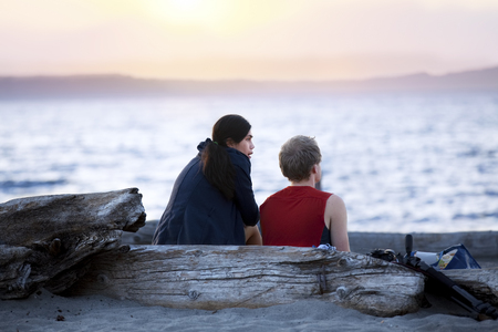 Young multiracial couple sitting  on driftwood log talking on beach at sunset. Diveristy, multiethnic relationship.の写真素材