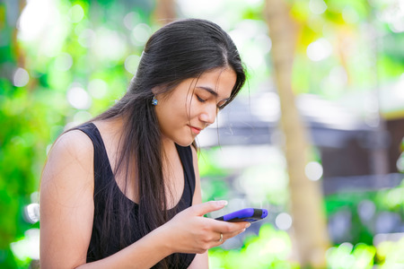Beautiful biracial Asian Caucasian teen girl talking on cell phone outdoors with green foliage in backgroundの写真素材