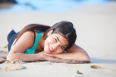 Beautiful biracial teen girl resting head on arms on sandy beach by ocean, wearing exercise outfitの写真素材