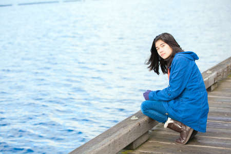 Biracial teen girl in blue jacket and boots with lonely or sad expression crouching down on wooden pier over water's edge, looking at cameraの写真素材