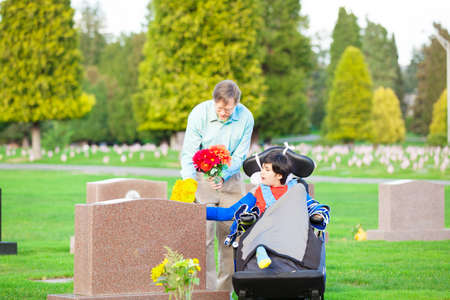 Father and disabled son in wheelchair holding flowers and visiting grave site in cemeteryの写真素材