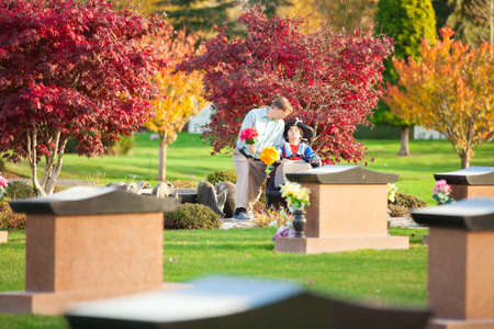 Father and disabled son in wheelchair holding flowers and visiting grave site in cemeteryの写真素材