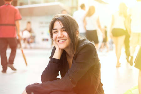 Beautiful biracial teen girl sitting by sidewalk, head in hand, smiling at camera with morning sunshine from the sideの写真素材