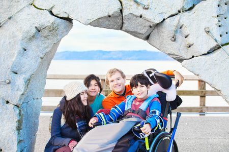 Smiling disabled ten year old  boy in wheelchair surrounded by friends and family outdoors near ocean water in background.の写真素材