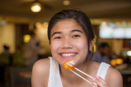 Biracial teen girl smiling, eating shrimp with chopsticks in restaurantの写真素材