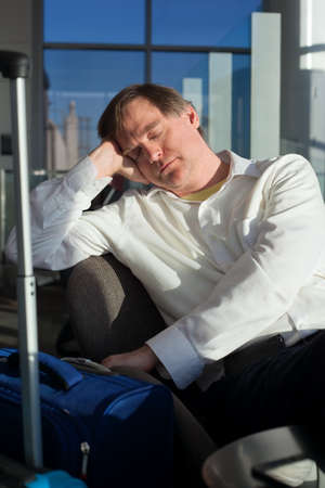 Tired Caucasian businessman sleeping at airport terminal next to luggage, sunlight streaming in through glass windowsの写真素材