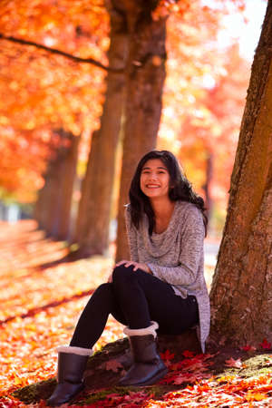 Biracial teen or young woman sitting under colorful maple leaves in autumn, with a carpet of fallen leaves underfoot.の写真素材