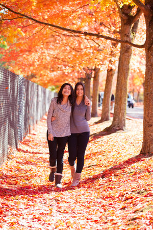 Two biracial teen girls walking together under bright red, orange and yellow maple tree, kicking...