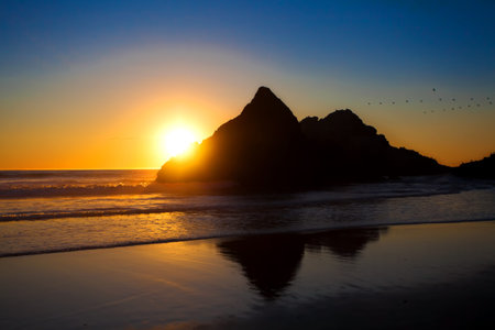 Beautiful yellow orange sunset peeking out from behind large rock along coast of Pacific Ocean...