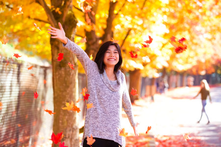 Biracial teen girl or young woman throwing autumn leaves into air, smiling