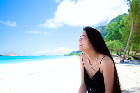 Side profile of smiling biracial teen girl or young woman with long black hair,  sitting at Hawaiian beach wearing black tank top の写真素材