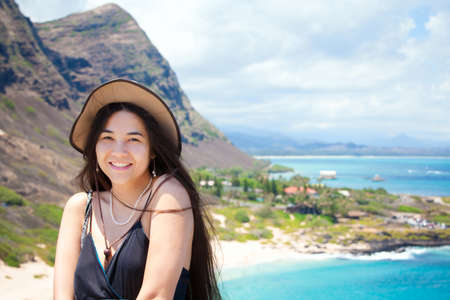 Smiling biracial teen girl, or young woman, smiling at camera with view of Hawaiian island in background from above looking down on  beach and water の写真素材