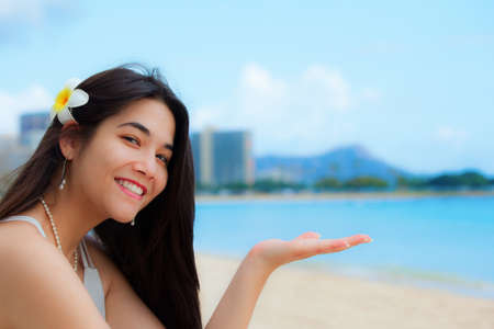 Smiling teen girl, or young woman, plumeria in long black hair, sitting on beach in Honolulu, Oahu with Diamond Head in the background. Hand palms up for product placementの写真素材
