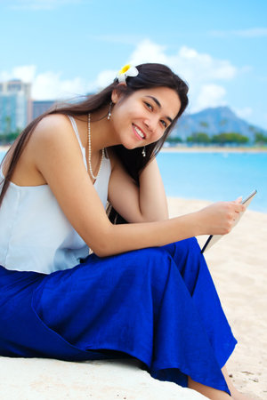 Smiling teen girl, or young woman, plumeria in long black hair, sitting on beach in Honolulu, Oahu holding computer tablet with Diamond Head in the backgroundの写真素材