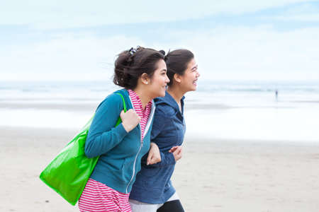 Two biracial Asian Caucasian  teenage girls talking while walking along beach on cool cloudy day の写真素材