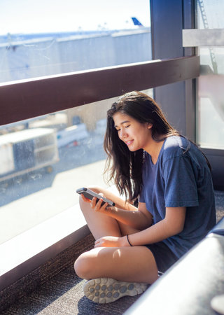 Biracial Asian, Caucasian teen girl sitting on floor at airport terminal waiting for flight while looking at smartphoneの写真素材