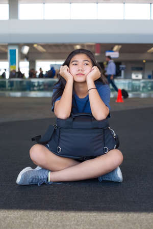 Biracial Asian Caucasian teen girl resting elbows on luggage while sitting on floor at airport, tiredの写真素材