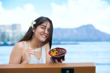 Smiling biracial Thai, Caucasian teen or young woman at Magic Lagoon eating Korean or Hawaiian BBQ rice bowl. Waikiki and Diamond Head in background.の写真素材