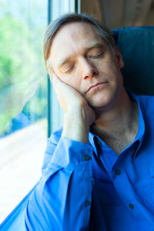 Handsome Caucasian man in early fifties resting head against train window, sleeping while commutingの写真素材