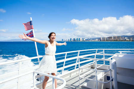 Biracial Asian, Caucasian teen girl standing on deck of cruise ship sailing in Waikiki bay,  arms outstretched in the wind. Honolulu Waikiki highrise hotels in background. の写真素材