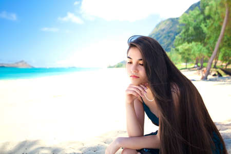 Young woman or teen girl sitting alone on beautiful Hawaiian beach looking sad or lonely, thinking, chin on handの写真素材