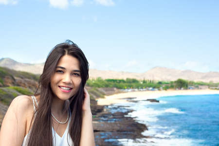 Biracial young Asian Caucasian woman or teen girl  smiling, standing overlooking  Hawaiian beach in backgroundの写真素材
