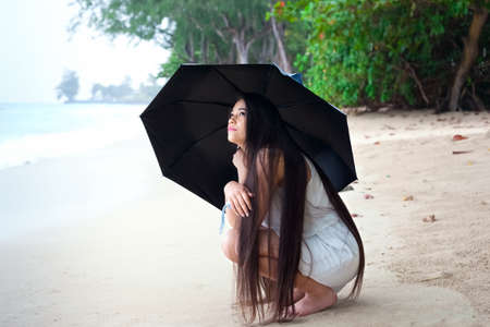 Young biracial Asian Caucasian woman or teen sitting on beach holding umbrella, looking  up checking for rain with sad or lonely expressionの写真素材