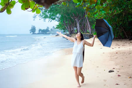 Young biracial Asian Caucasian woman or teen walking on beach holding umbrella on overcast rainy day, arms outstreched enjoying the rain, smilingの写真素材