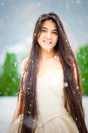 Smiling biracial Asian Caucasian young woman in gold dress standing outdoors with snow falling の写真素材
