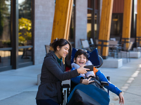 Disabled eleven year old biracial boy in wheelchair watching video on smartphone with his two...