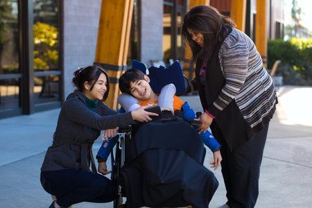 Disabled eleven year old biracial boy in wheelchair watching video on smartphone with his two caregivers outdoorsの写真素材