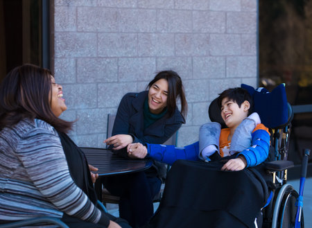 Eleven year old disabled biracial Asian Caucasian boy in wheelchair outdoors, sitting and talking with his two multiethnic caregiversの写真素材