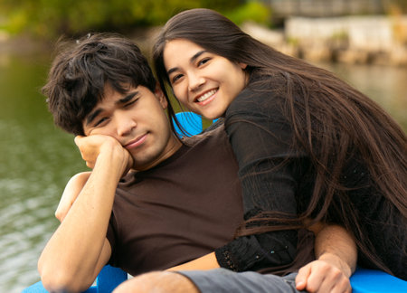 Biracial teen siblings outdoors relaxing in blue adirondack chairs by lake on wooden pier