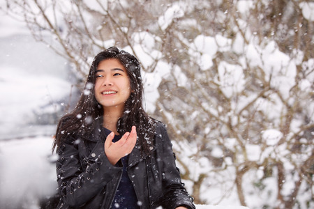 Biracial teen girl wearing black leather jacket standing outside in winter during heavy snowfall, enjoying the weatherの写真素材