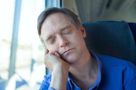 Caucasian man wearing blue shirt in early fifties sleeping and resting head on hand near train windowの写真素材