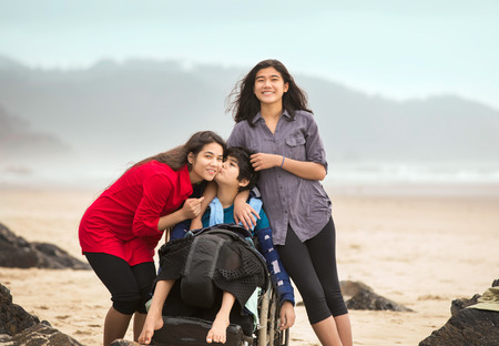 Disabled younger brother in wheelchair giving older sister a kiss while they stand together by ocean on misty foggy beachの写真素材