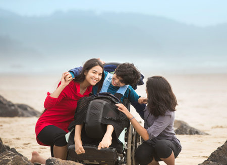 Disabled younger brother in wheelchair laughing and giving older sistesr a hug while they sit...