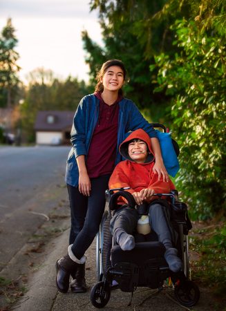 Big sister pushing disabled little brother in wheelchair around neighborhood, laughing and smilingの写真素材