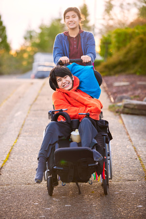 Big sister pushing disabled little brother in wheelchair around neighborhood, laughing and smilingの写真素材