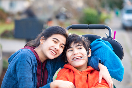 Biracial big sister lovingly hugging disabled little brother in wheelchairoutdoors, smilingの写真素材