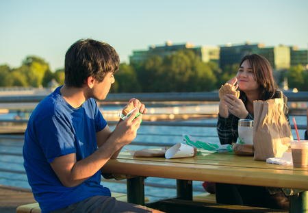 Two young biracial Asian, Caucasian adults sitting outdoors by lake in urban area eating hamburgers and talking together at picnic table near sunset の写真素材