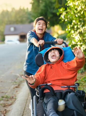 Teen girl running and smiling while pushing disabled little boy in wheelchair outdoors の写真素材