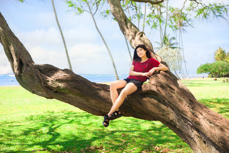 Biracial Asian teen girl in red shirt and shorts sitting on large tree branch at empty Kawaikui...