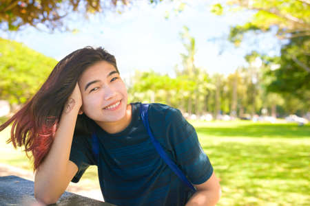 Smiling biracial Asian Caucasian teen girl or young woman sitting at table outdoors in park on sunny day, leaning head on hand and looking at camera, happyの写真素材