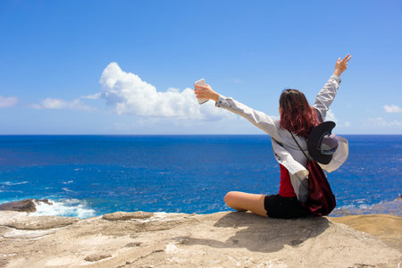 Biracial Asian Caucasian teen girl sittig on rock edge of cliff over blue Hawaiian ocean on sunny...
