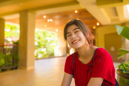 Smiling biracial Asian Caucasian teen girl or young woman sitting in empty outdoor open air lobby in tropical sunny Hawaii, wearinng red shirtの写真素材